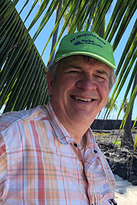 David Legler smiles at the camera with a blue sky and palm leaves in the background.