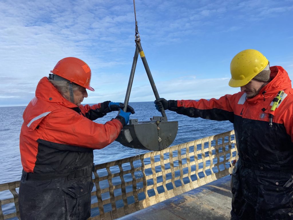 Lee Cooper and graduate student Brian Marx on the 2023 NOAA Arctic Marine Ecosystems Cruise hold a Van Veen Grab sampler filled with a fresh sample of seafloor sediment and the life found within the sediment.