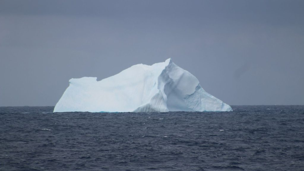 A lone iceberg floats in the open ocean with a gray, overcast sky in the background. 