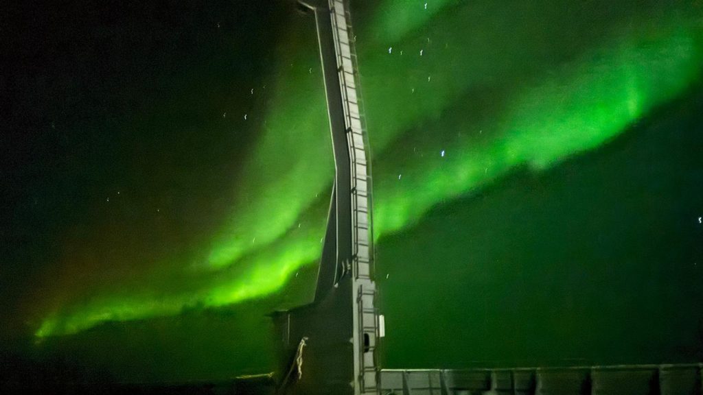 The Aurora Australis paints the sky green with the ship in the foreground.