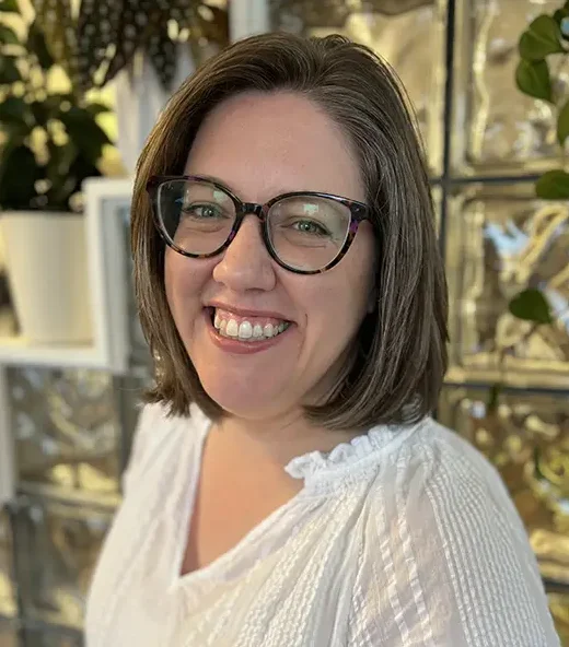 Heather, a woman with straight, shoulder-length hair and glasses in a white blouse smiles at the camera.