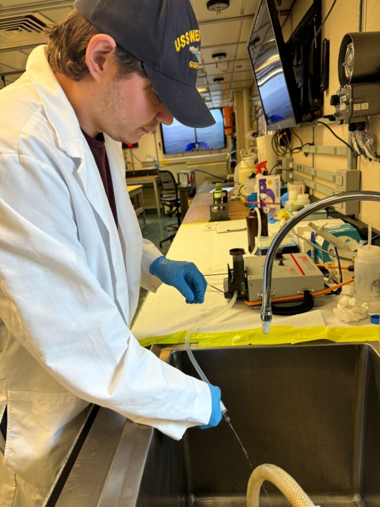 Robert, wearing a blue baseball cap, white lab coat, and blue nylon gloves, filters water for environmental DNA sampling.